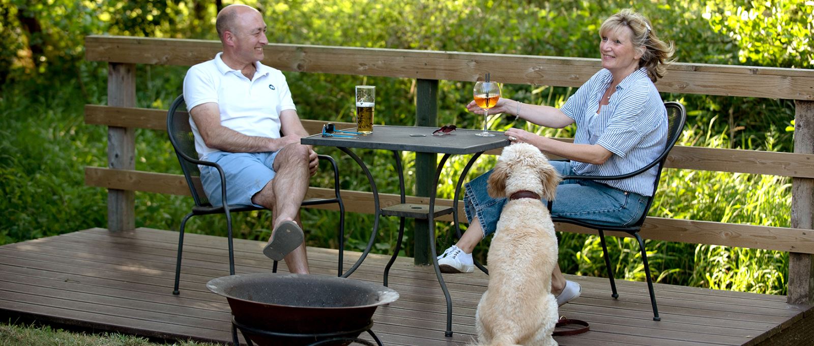A couple and a dog sit on a decking area of a glamping unit at Back of Beyond
